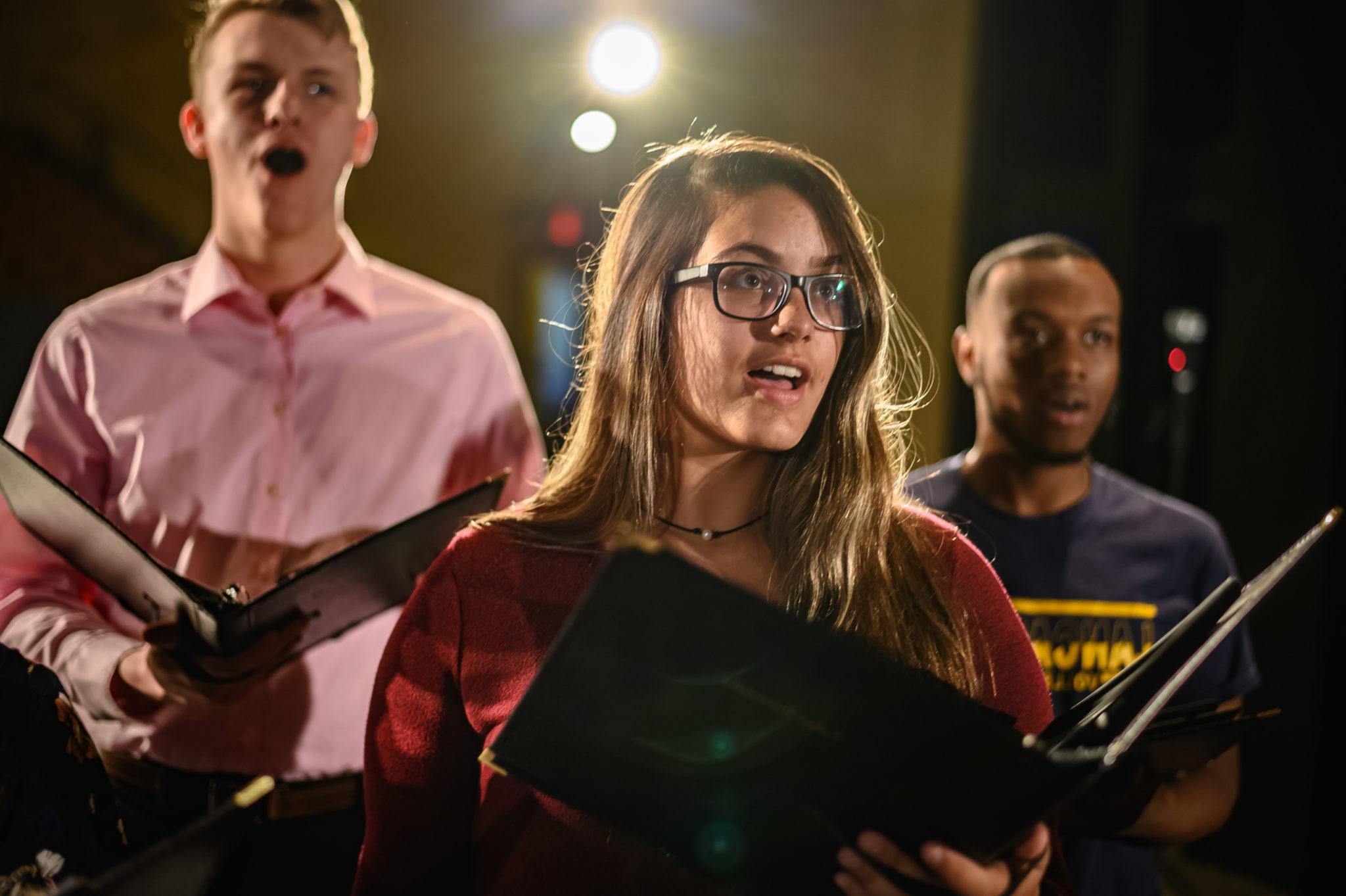 Three singers holding folders, close to one another, on stage